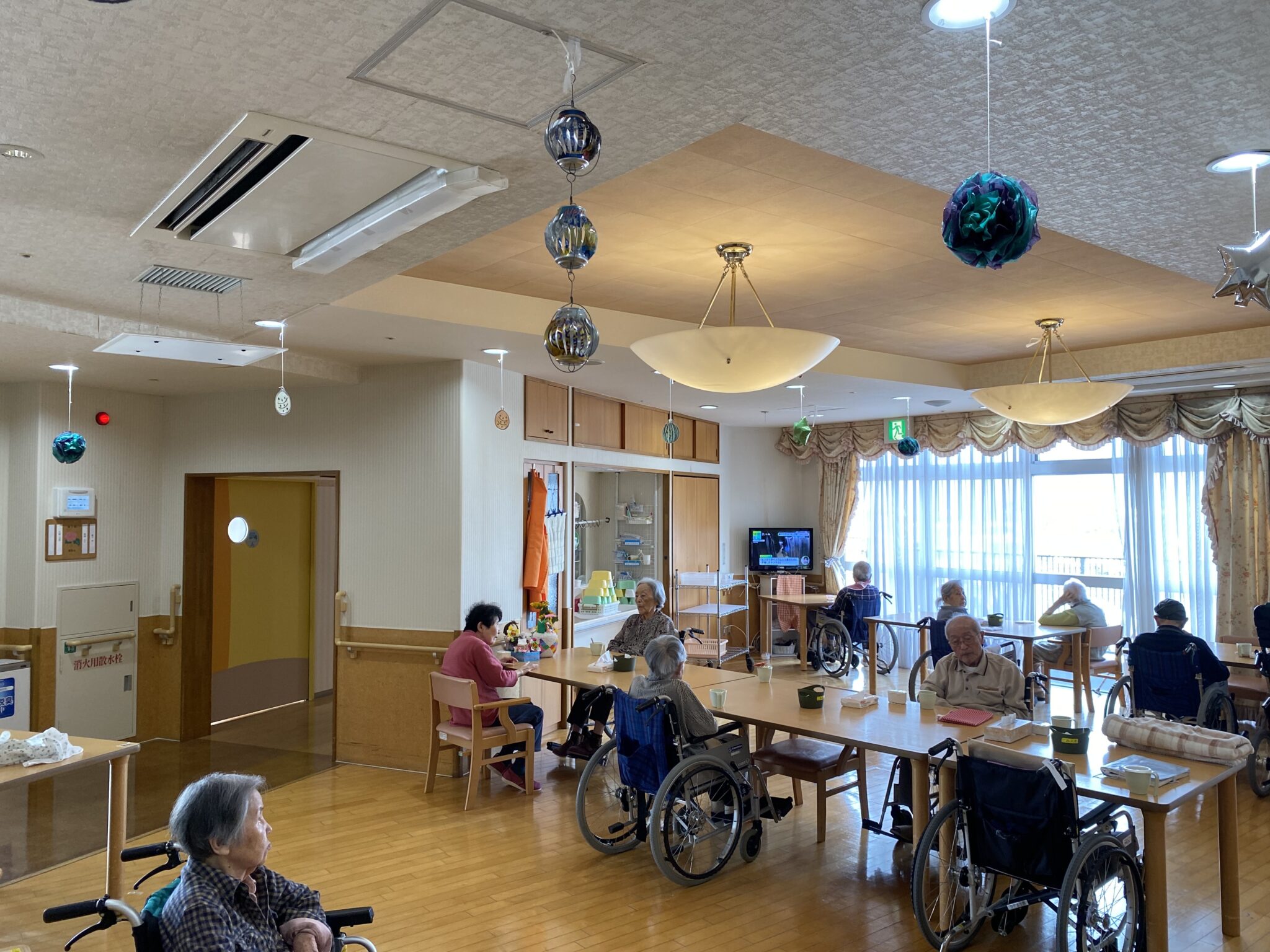 介護施設の共用リビング。複数の入居者が車いすを使用しながら過ごす明るい空間。